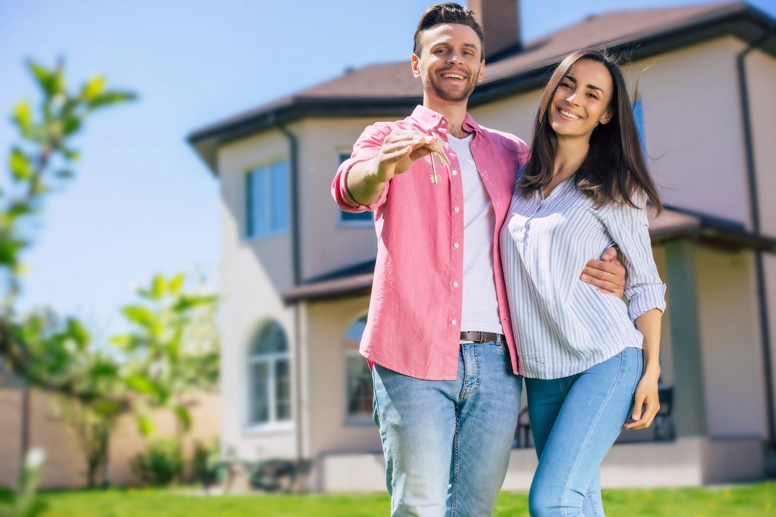 Modern beautiful excited young couple in love standing in front of the new big house with the keys in hands and hugging while they celebrating this buying