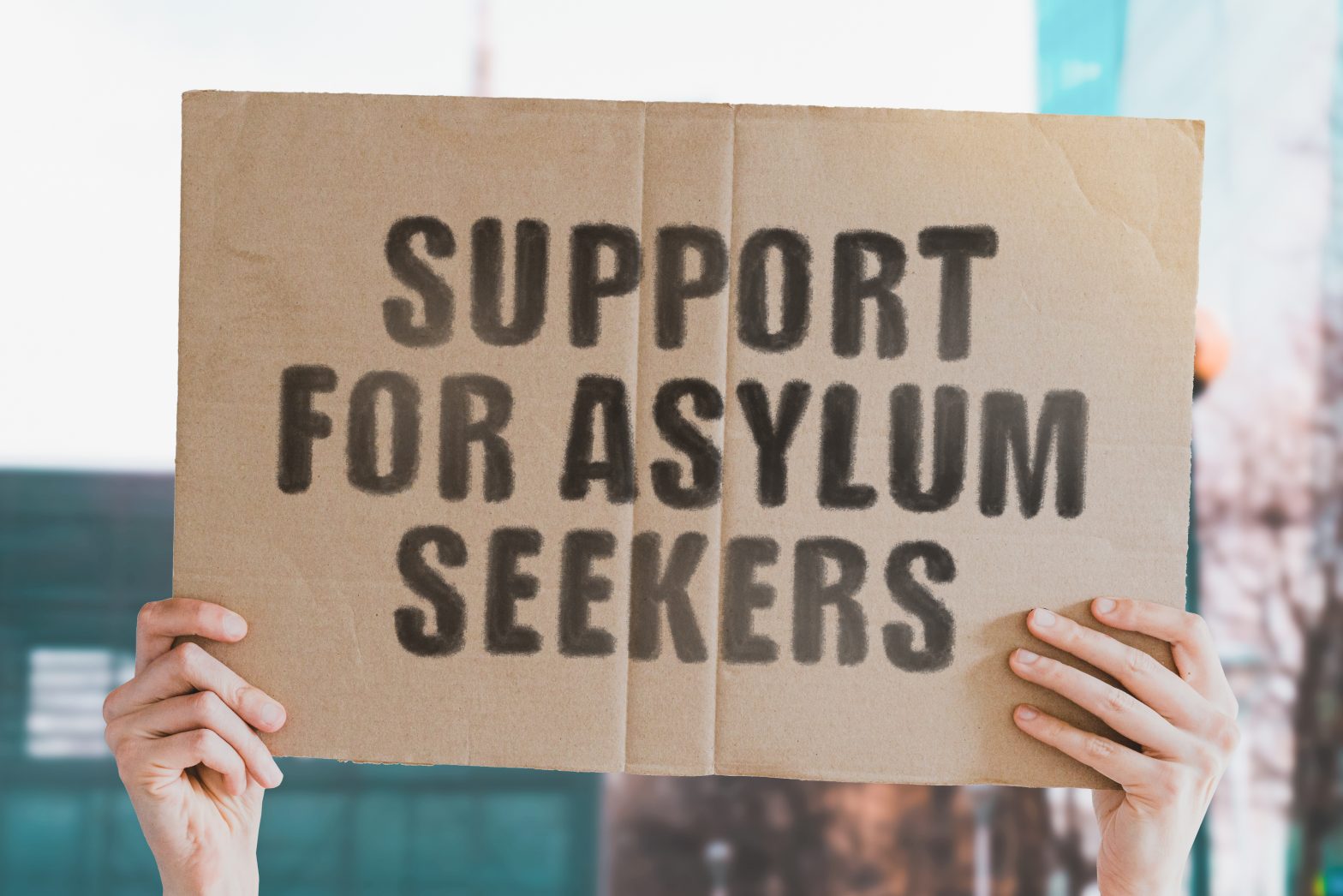 The phrase " Support for asylum seekers " on a banner in men's hand with blurred background. Immigrants. Refugees. Immigration. Charity. Help. Human rights. Shelter. Donation
