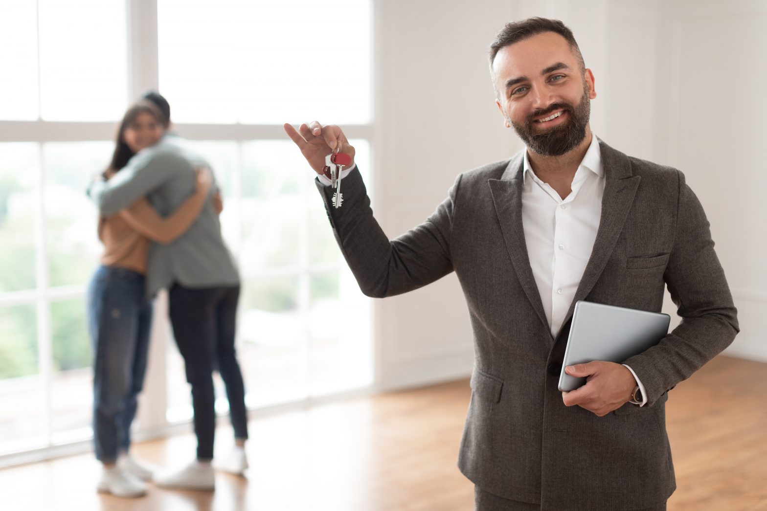 House Ownership. Portrait Of Smiling Professional Real Estate Agent In Suit Holding And Showing Key To Camera, Selective Focus. Happy Spouses Embracing Standing In New Home In Blurred Background
