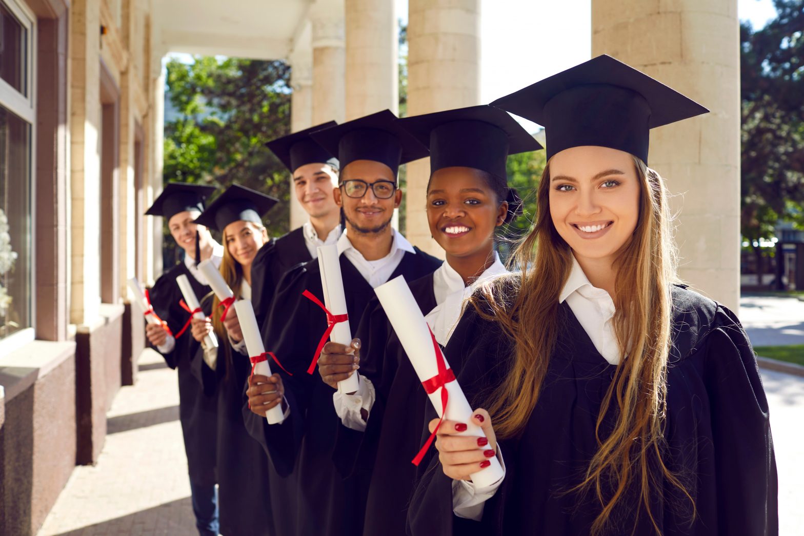 Multinational group of attractive students-graduate in mortar boards cap and ceremonial robes holding diploma in their hands, look very positive and happy, smiling snow-white smile at the camera.