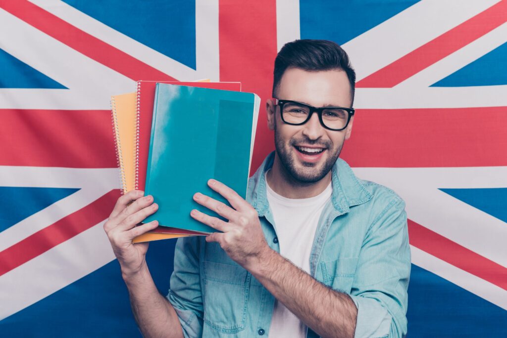 English language learning concept-portrait of cheerful attractive man with bristle showing colorful copy books standing over English flag background