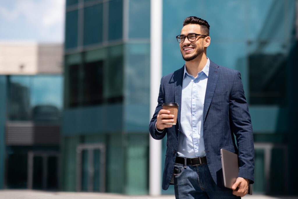 Cheerful middle eastern young businessman in glasses with laptop and coffee to go walking by street towards modern office building, going to job in the morning, panorama with copy space
