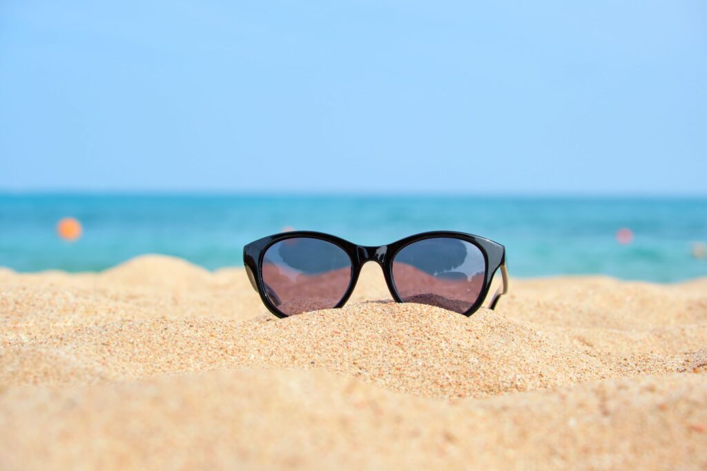 Closeup of black protective sunglasses on sandy beach at tropical seaside on warm sunny day. Summer vacation concept.