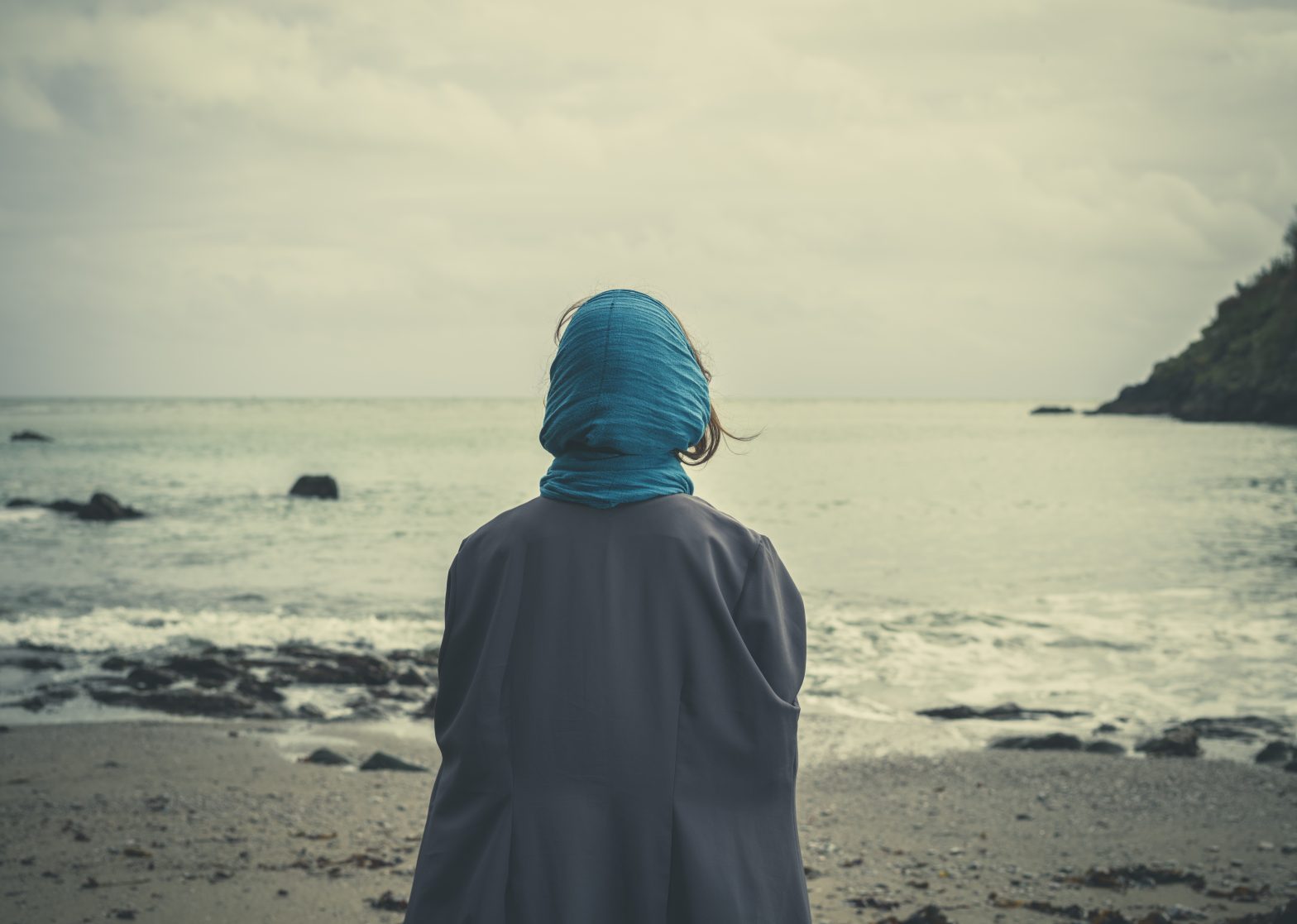 A young woman swearing a headscarf is standing on the beach and is looking out to sea on a windy and cold day