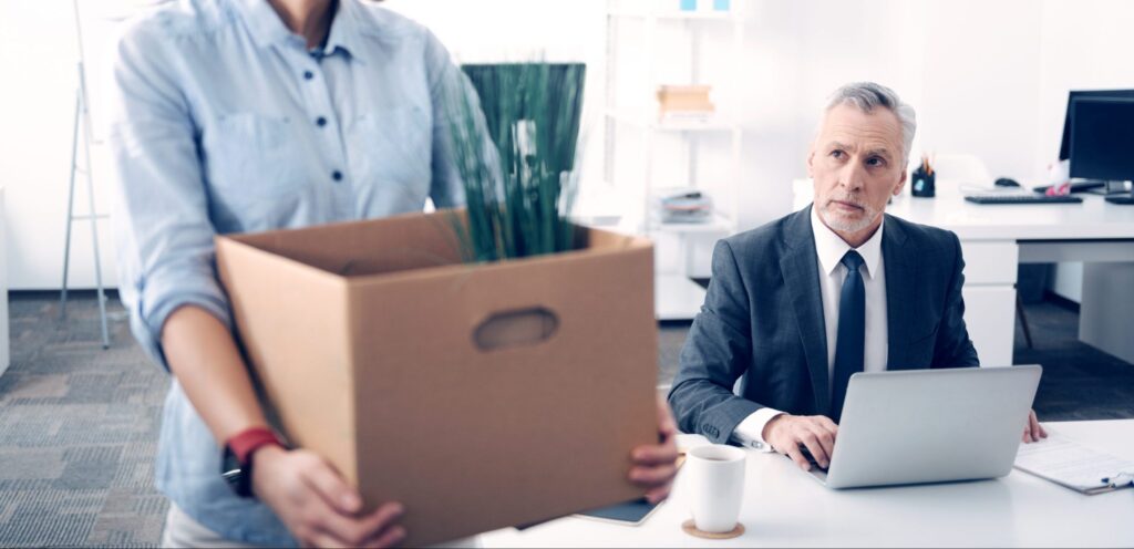 Serious chief working on a laptop and looking at his former office worker leaving the office with a box full of different supplies.