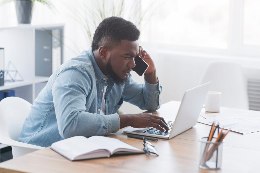 Worried black businessman talking on cellphone and looking at laptop screen, trying to solve problems online, side view with copy space