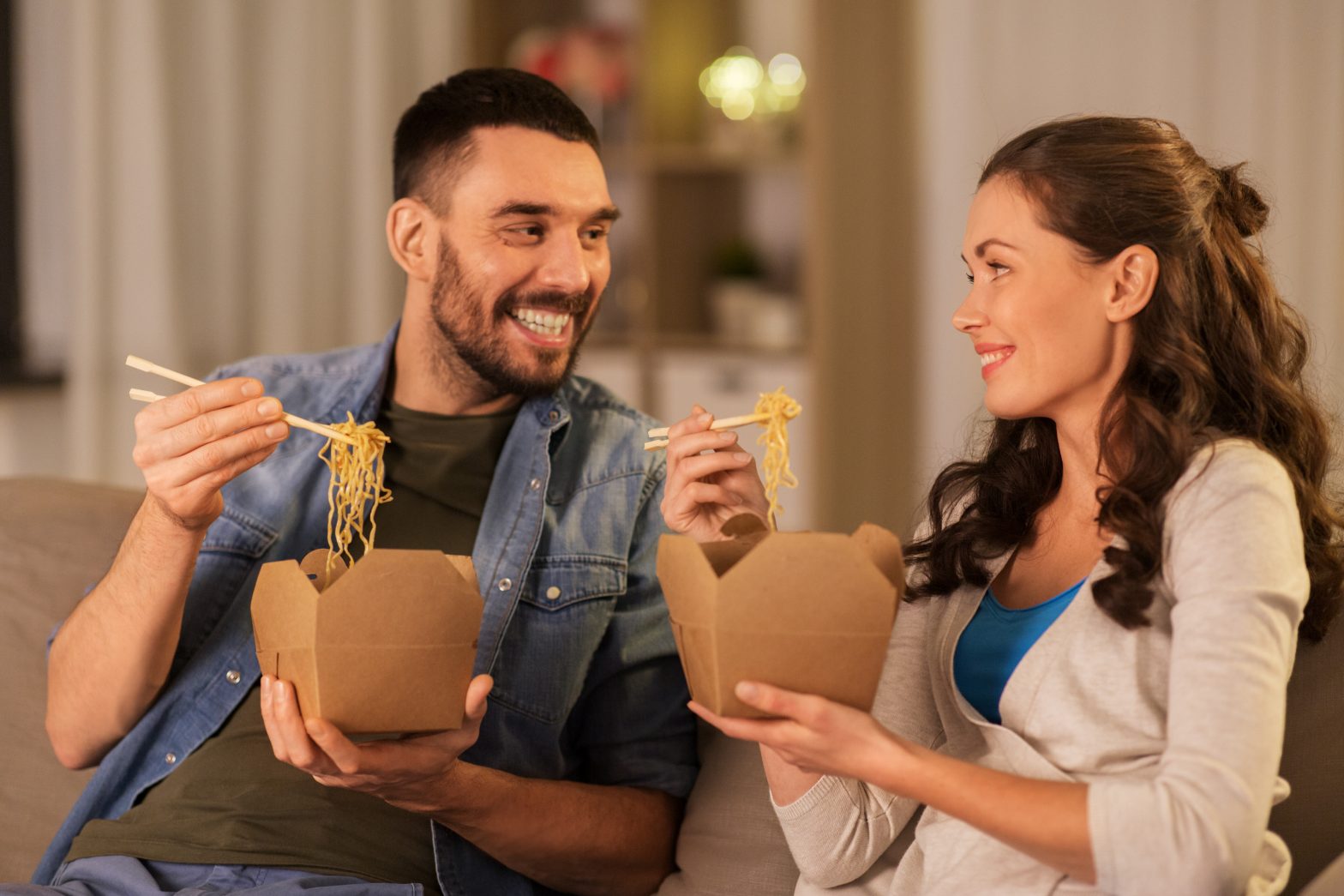 fast food and people concept - happy couple eating takeaway noodles with chopstick at home in evening