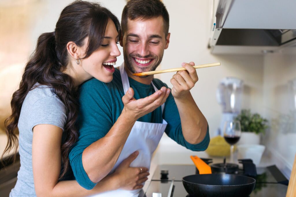 Shot of handsome young man giving his wife to try the food he is preparing in the kitchen at home.