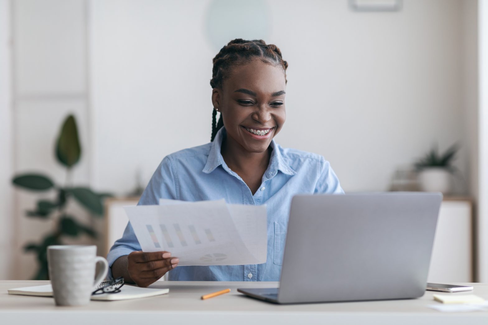 Cheerful black millennial businesswoman working with laptop and documents in modern office, checking financial reports, free space