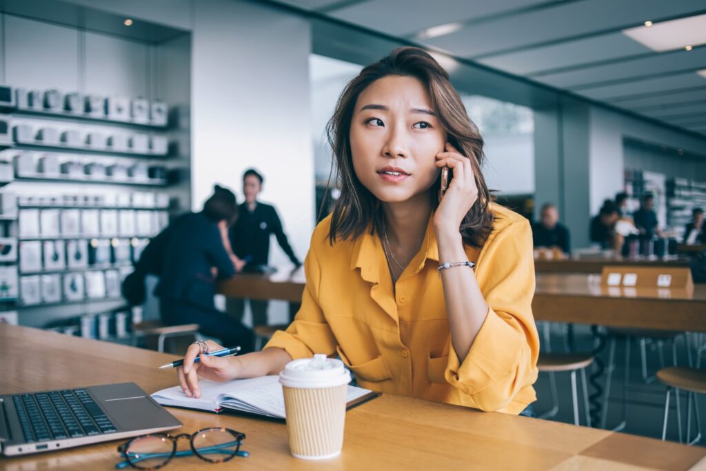 Concerned Asian female speaking on phone and taking notes at table with laptop and glasses in workspace in Hong Kong city