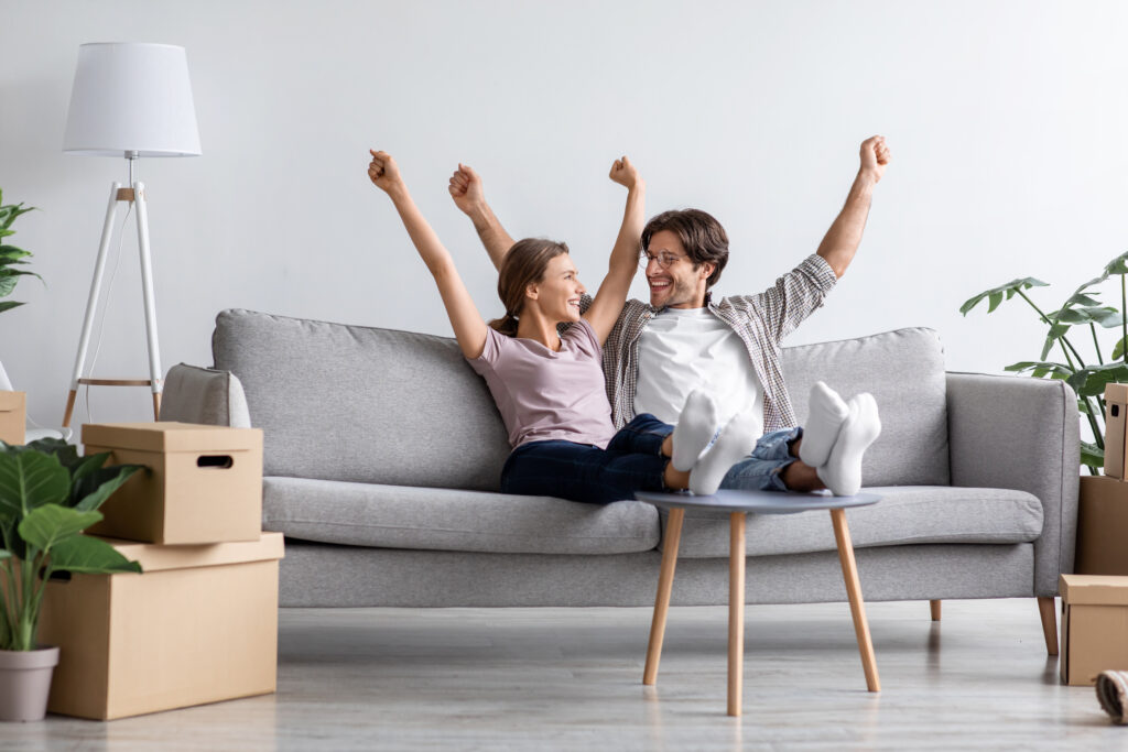 Happy excited european young husband and wife rejoice raise their hands up on sofa in living room interior