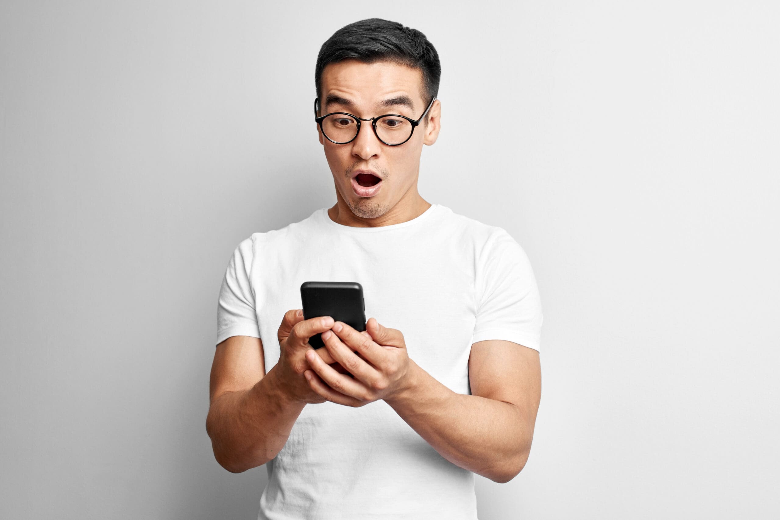 Asian guy in white studio looks surprised at screen of mobile phone