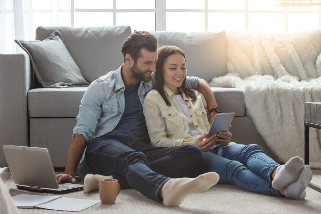 Couple sitting on a gray sofa, sharing a tablet while a laptop sits on the floor nearby in a bright living room.