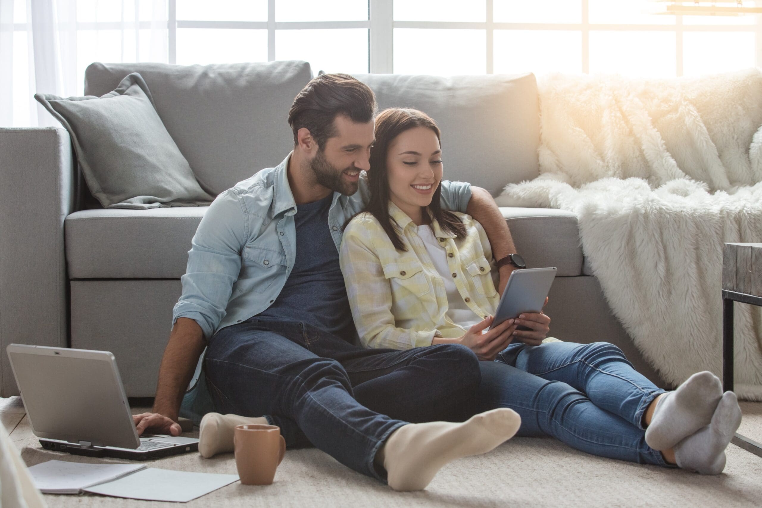 Couple sitting on a gray sofa, sharing a tablet while a laptop sits on the floor nearby in a bright living room.