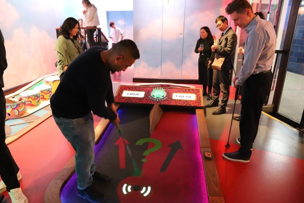 IMG_4478 - Lisa's Law Solicitors - Lisa's Law Solicitors Man in a dark shirt bends to play a tabletop game labeled TRUE/FALSE while a group of adults watches in a colorful arcade-like room with drinks in hand.