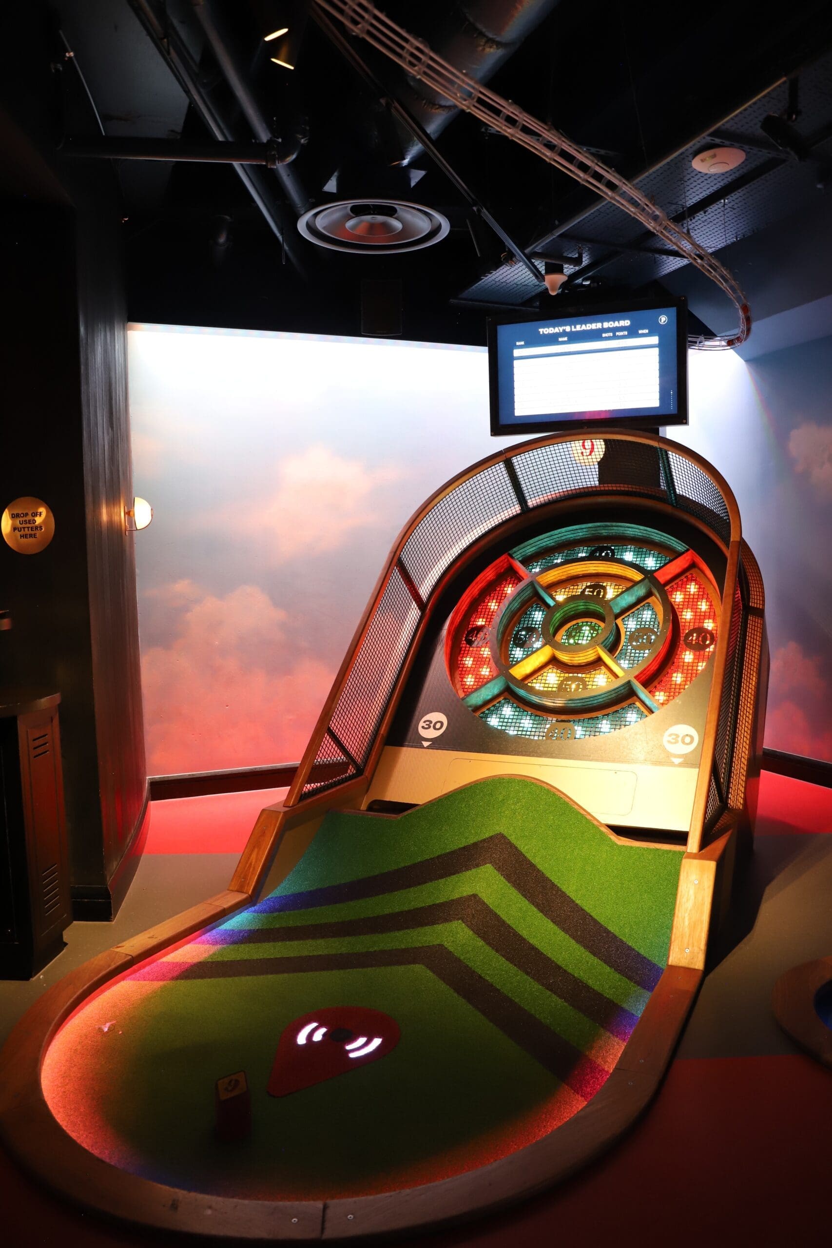 Indoor arcade scene with a large colorfully lit circular wheel target, green carpet, and a digital leaderboard hanging above the game area on a dark ceiling.