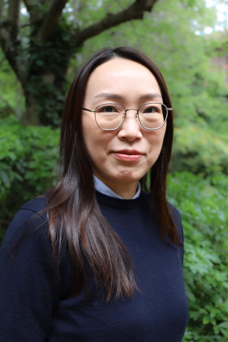 Close-up portrait of a woman wearing round glasses, outdoors with green foliage in the background.