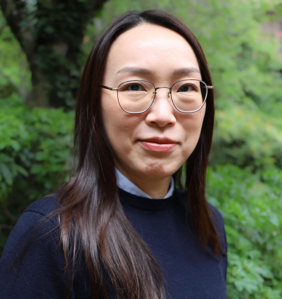 Close-up portrait of a woman wearing round glasses, outdoors with green foliage in the background.