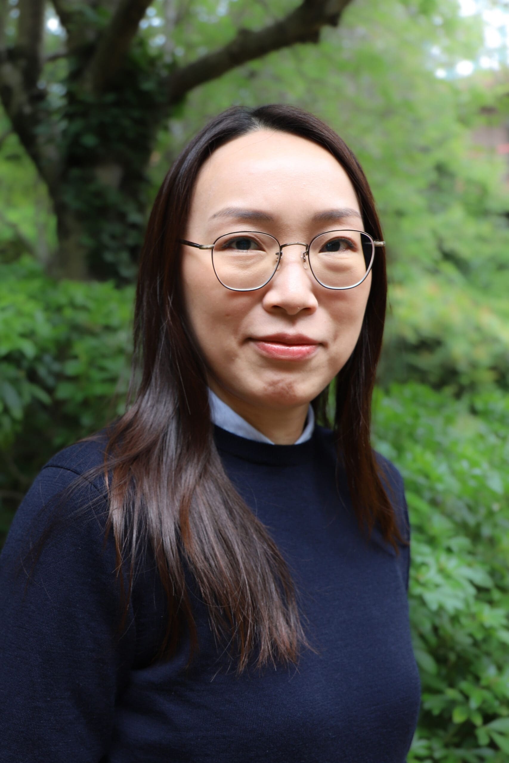 Close-up portrait of a woman wearing round glasses, outdoors with green foliage in the background.