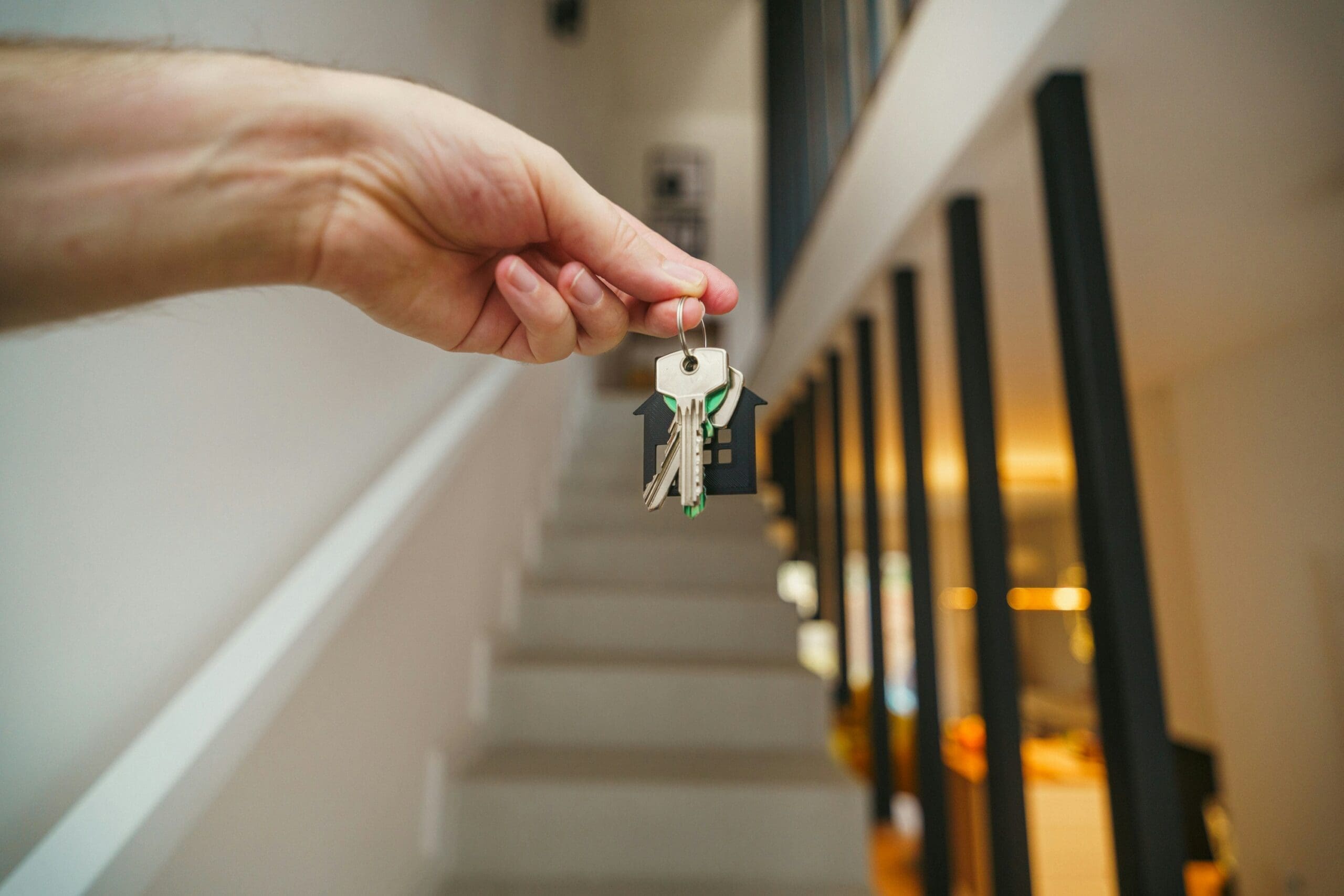 Person's hand holds a keyring with several keys in focus, a hallway railing blurred in the background.
