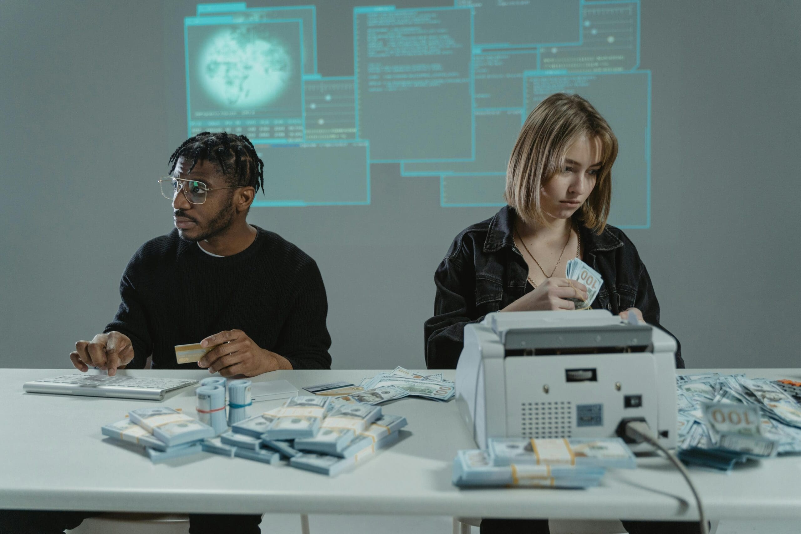 Two people sit at a table surrounded by large piles of cash, with a printer and calculator in view and holographic screens projected on the wall behind them.