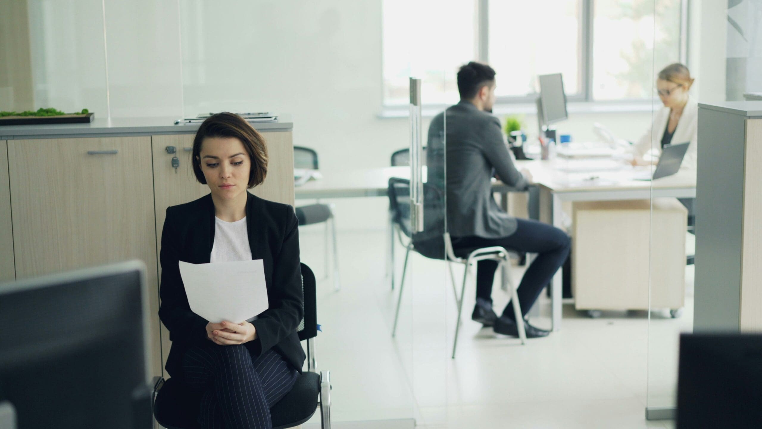 Young woman waiting in modern office reading her CV while another candidate is talking to interviewer then girl is entering room and greeting manager.