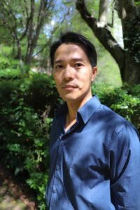 Close-up portrait of a man wearing a blue shirt standing in a sunlit green garden with trees and plants in the background.