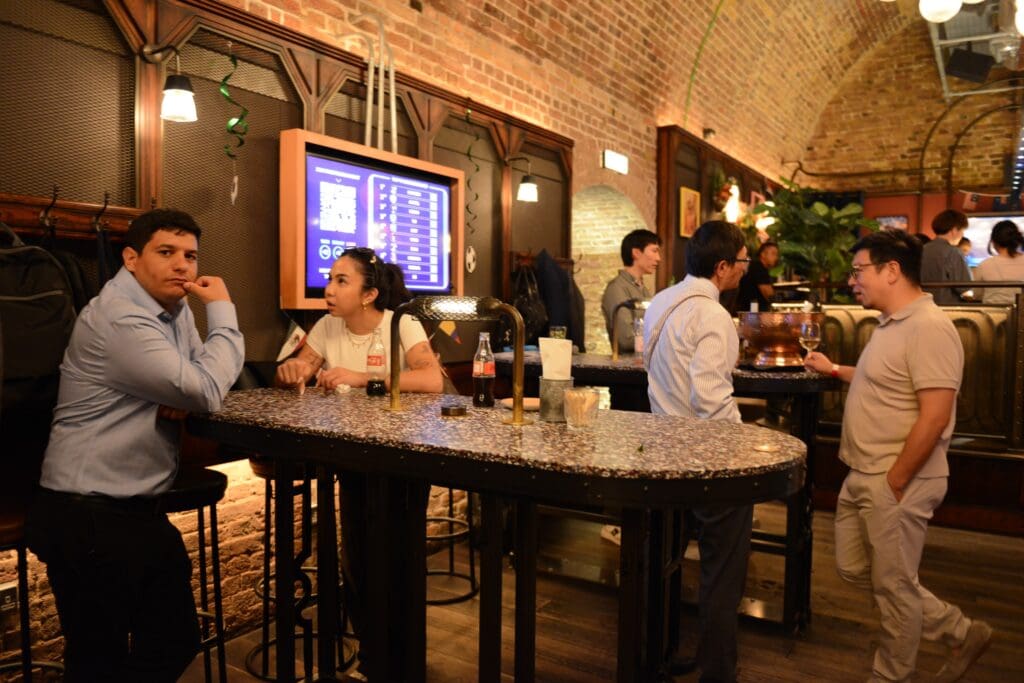 DSC_9232 - Lisa's Law Solicitors Patrons sit at a high, speckled bar counter in a brick-walled venue, chatting with a digital menu screen glowing behind them.