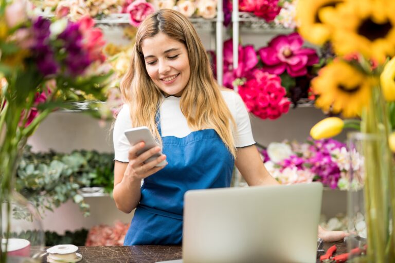 Smiling flower shop worker in a blue apron checks her phone among colorful blooms.
