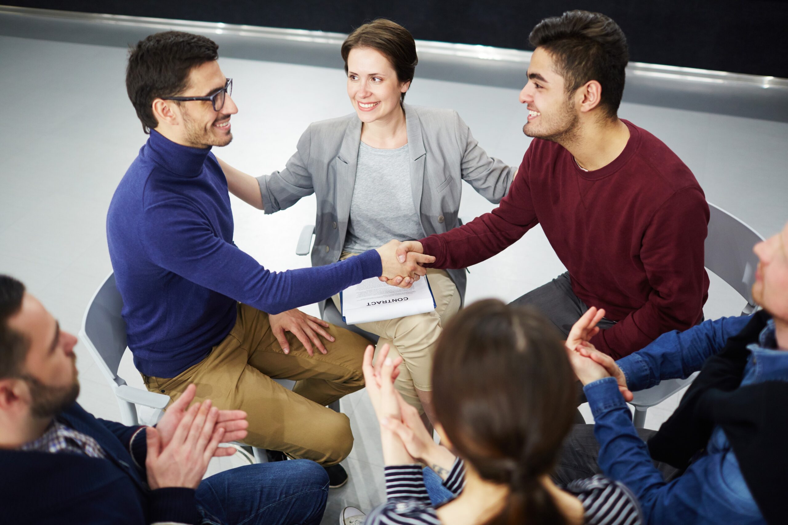 People seated in a circle, two men shake hands while others applaud and smile nearby.