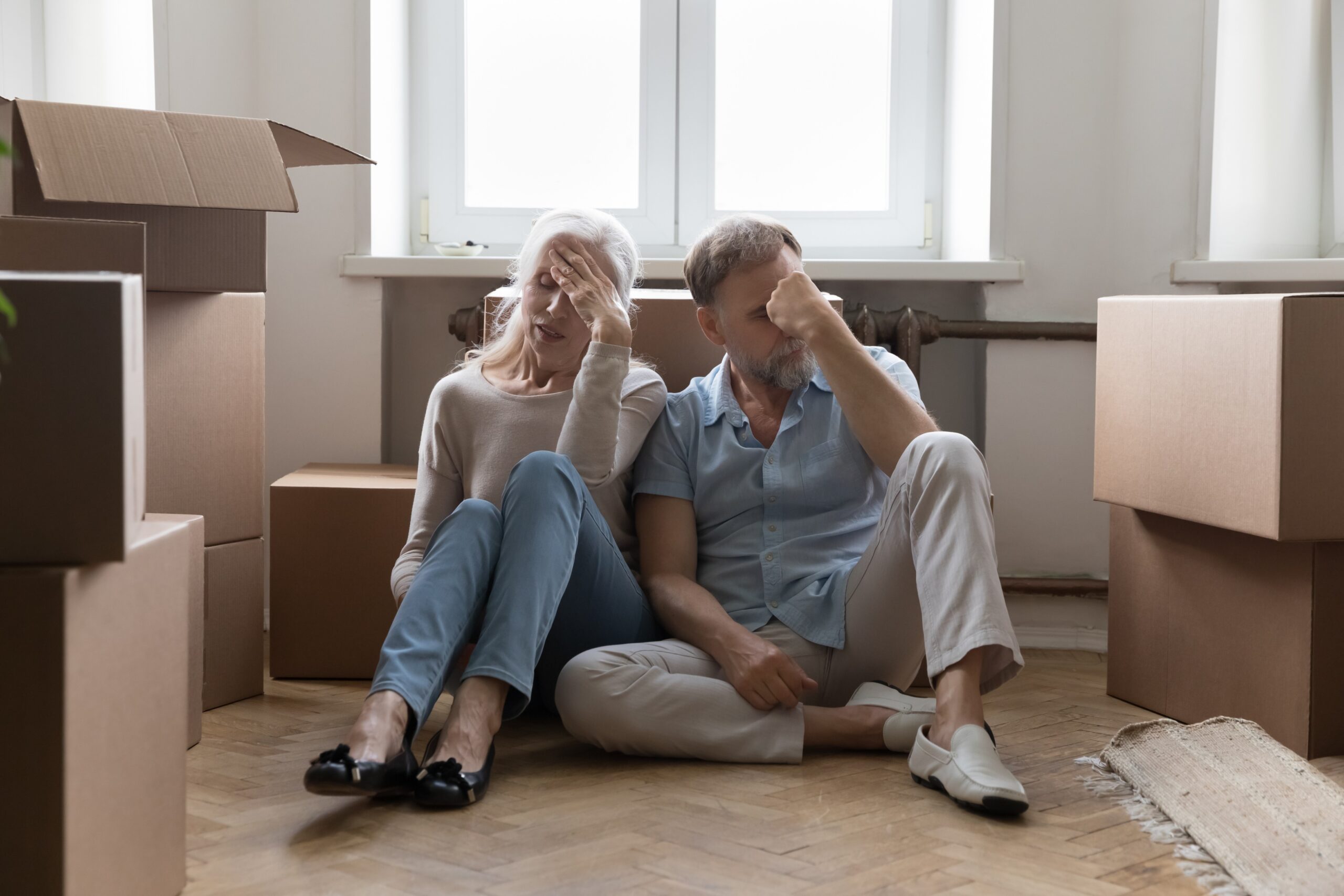 Elder couple of homeowners exhausted with relocation, packing, unpacking cardboard boxes, sitting on floor of new home at paper containers.