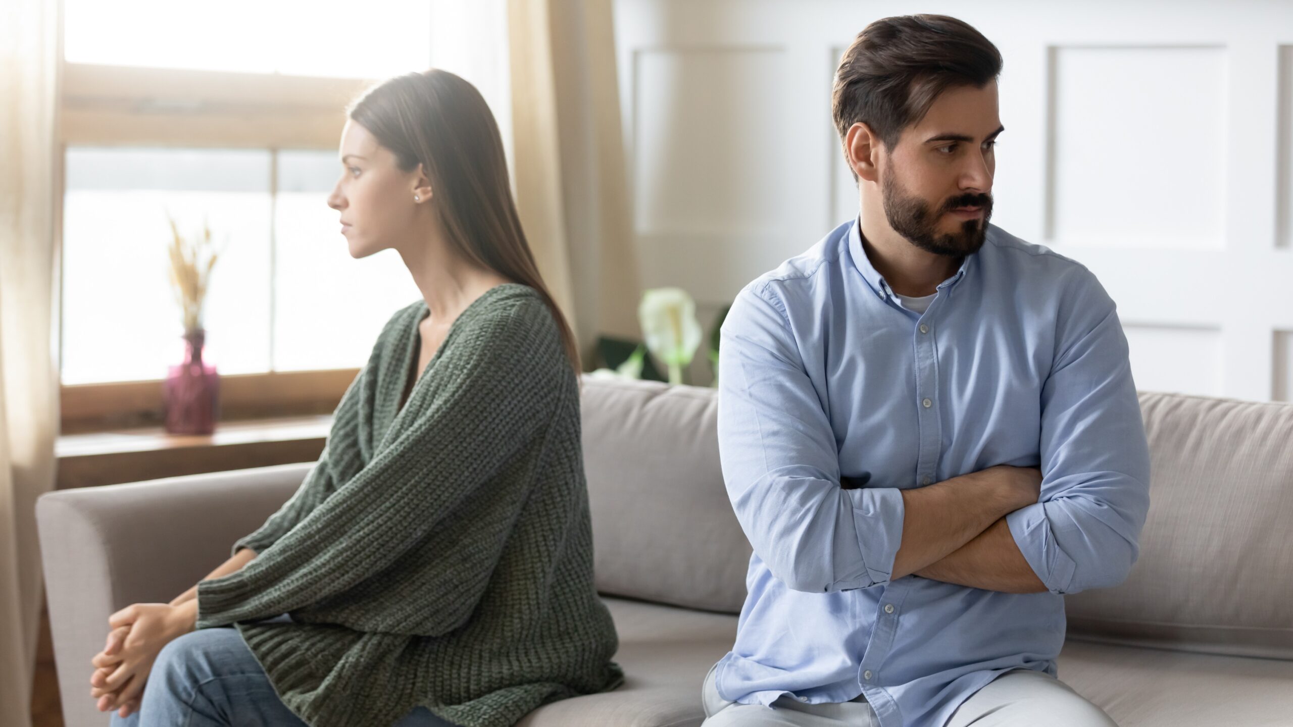 Angry young Caucasian couple sit separate on couch ignore avoid talking after family fight misunderstanding.