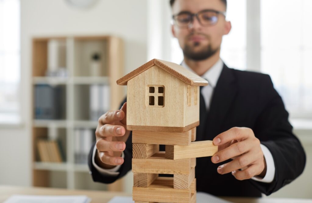Male entrepreneur removes wooden blocks from tower foundation of little miniature toy house model as symbol of business risks