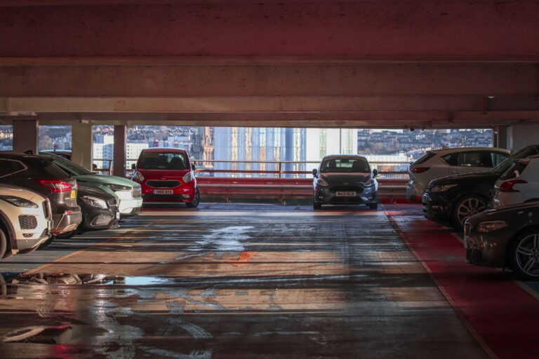 Interior of a parking garage with rows of cars on either side and a city skyline visible through the open end.